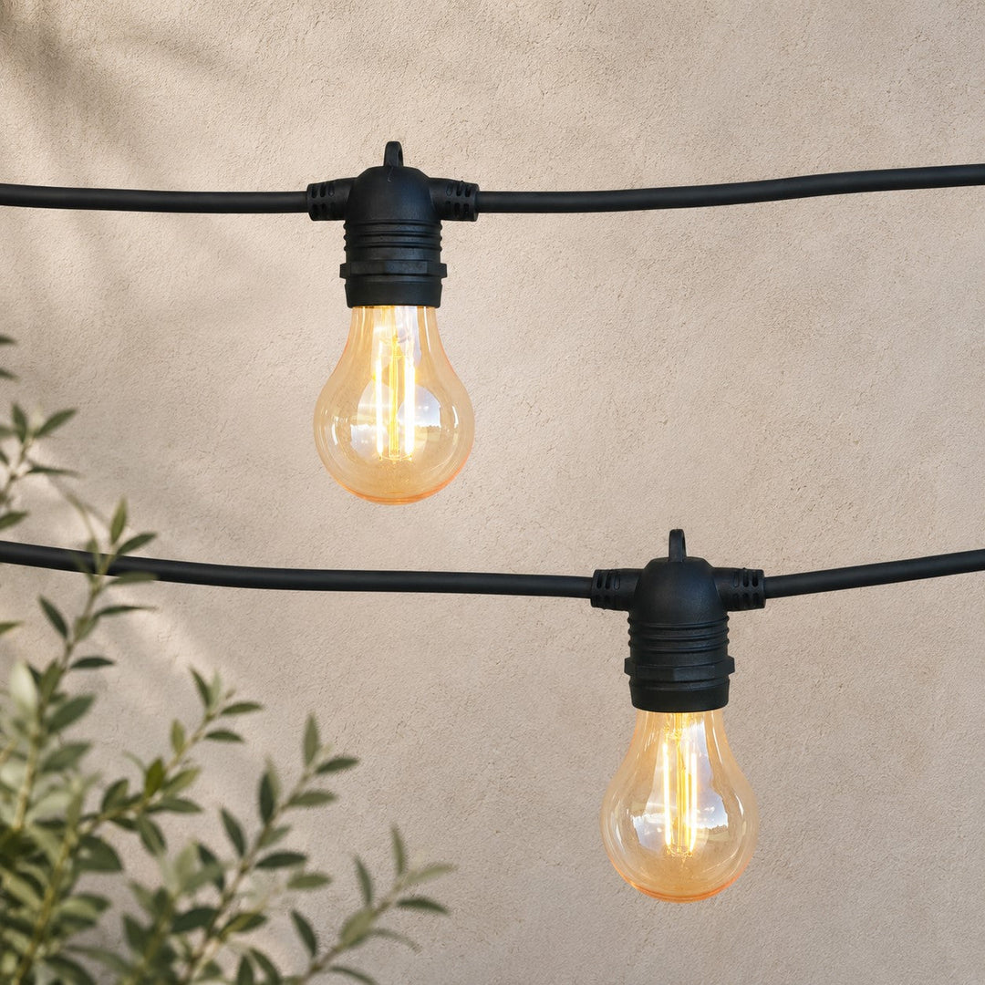 Black outdoor string lights with clear glass bulbs glowing warm yellow, suspended against a beige stucco wall with green leaves in the lower left corner.