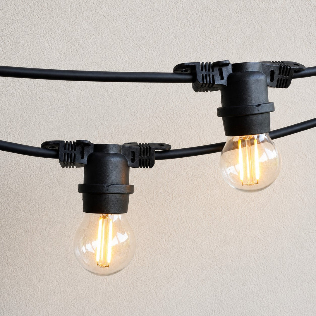 Black outdoor string lights with clear glass bulbs, shown illuminated against a light beige textured wall.