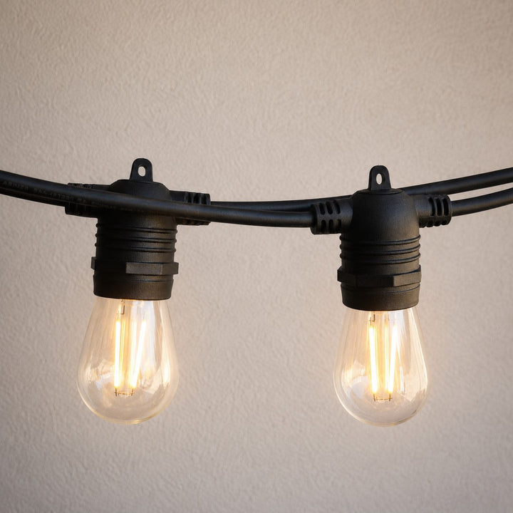Black string light bulbs with clear glass and visible warm glowing filaments, mounted on a black cable against a light beige textured wall.